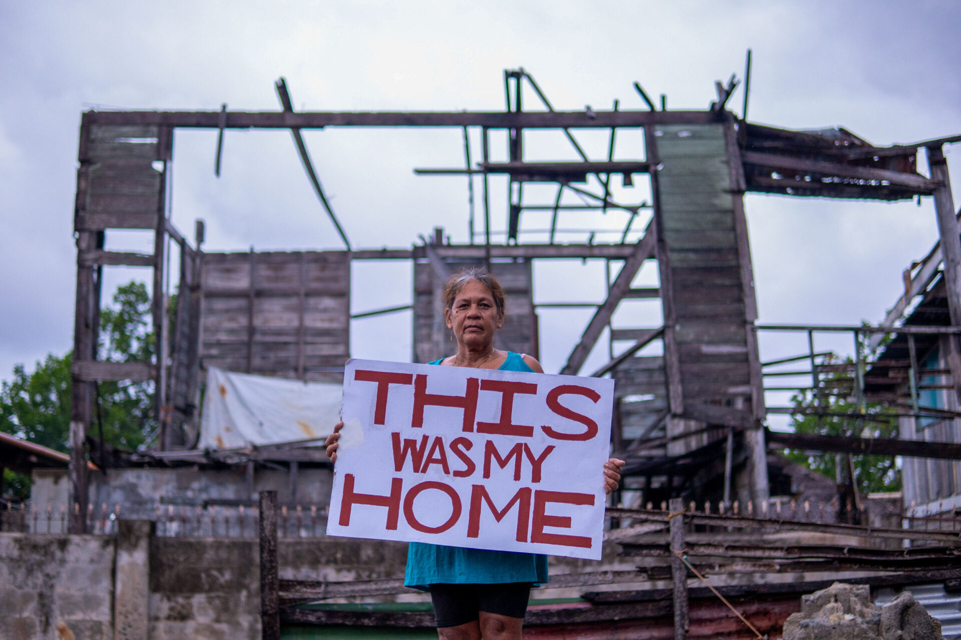 A woman holds up a sign saying "This was my Home" in front of her destroyed house.