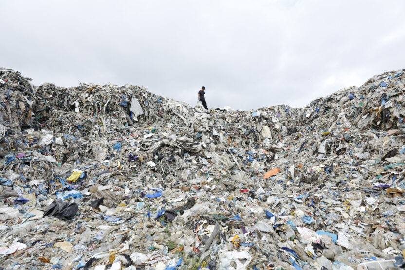 A huge heap of waste can be seen in the foreground with a tiny person standing in the background on top.
