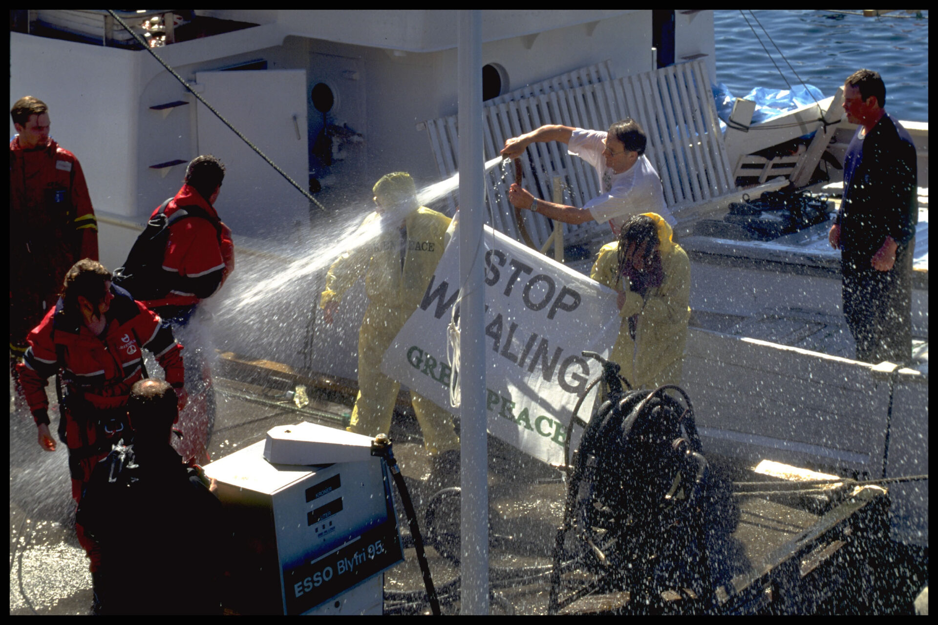Greenpeace activists wearing yellow overalls are hosed with water when trying to block a whaling ship in Norway. They hold a banner that says 'Stop Whaling'.