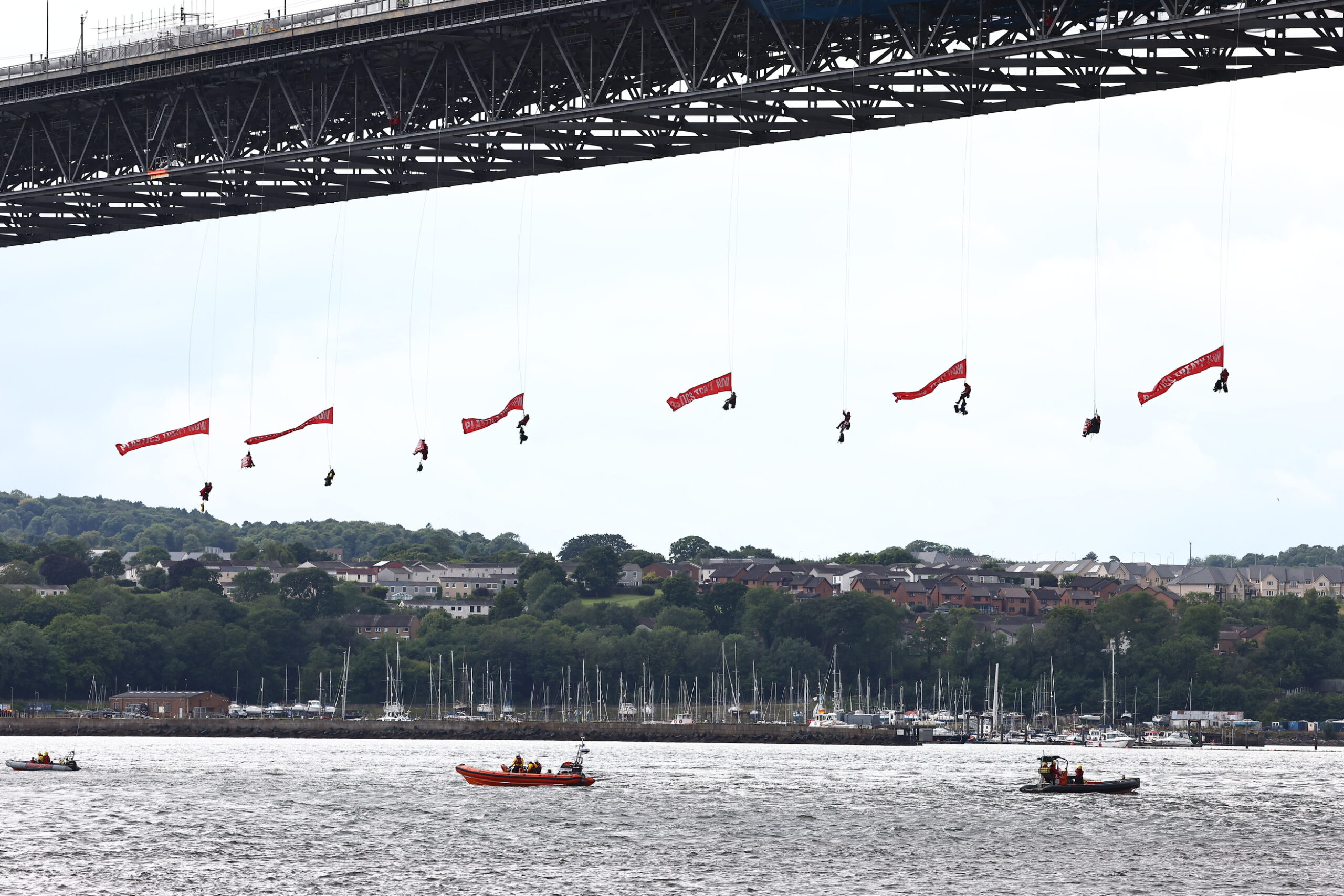 Greenpeace activists hang on long ropes from the Forth Bridge, with red banners streaming in the wind.