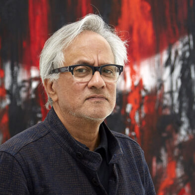 Headshot of Anish Kapoor in front of a black and red canvas.
