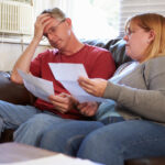 A man and woman sit on a sofa, looking worried as they examine household bills.