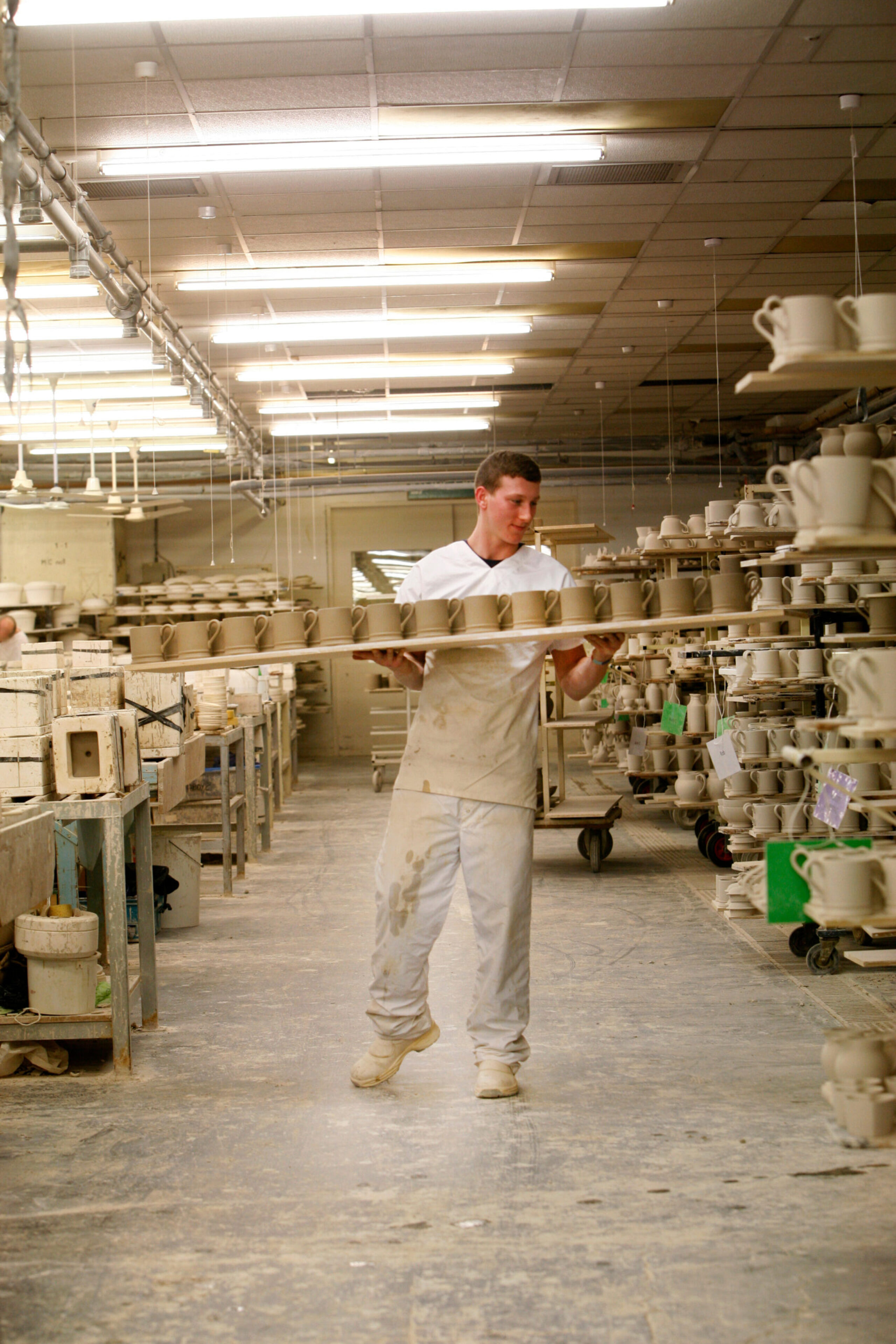 A worker in white overalls carries a tray of unfinished mugs through a ceramics factory.