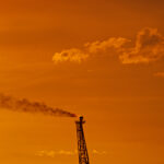 An oil refinery flare stack emits smoke against a dramatic orange sunset sky.