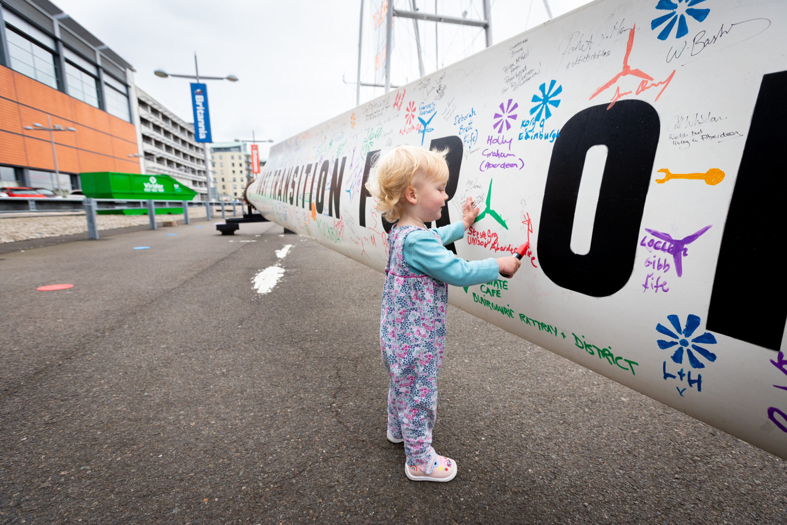 A young child draws on a white turbine blade covered in colourful handwritten messages.