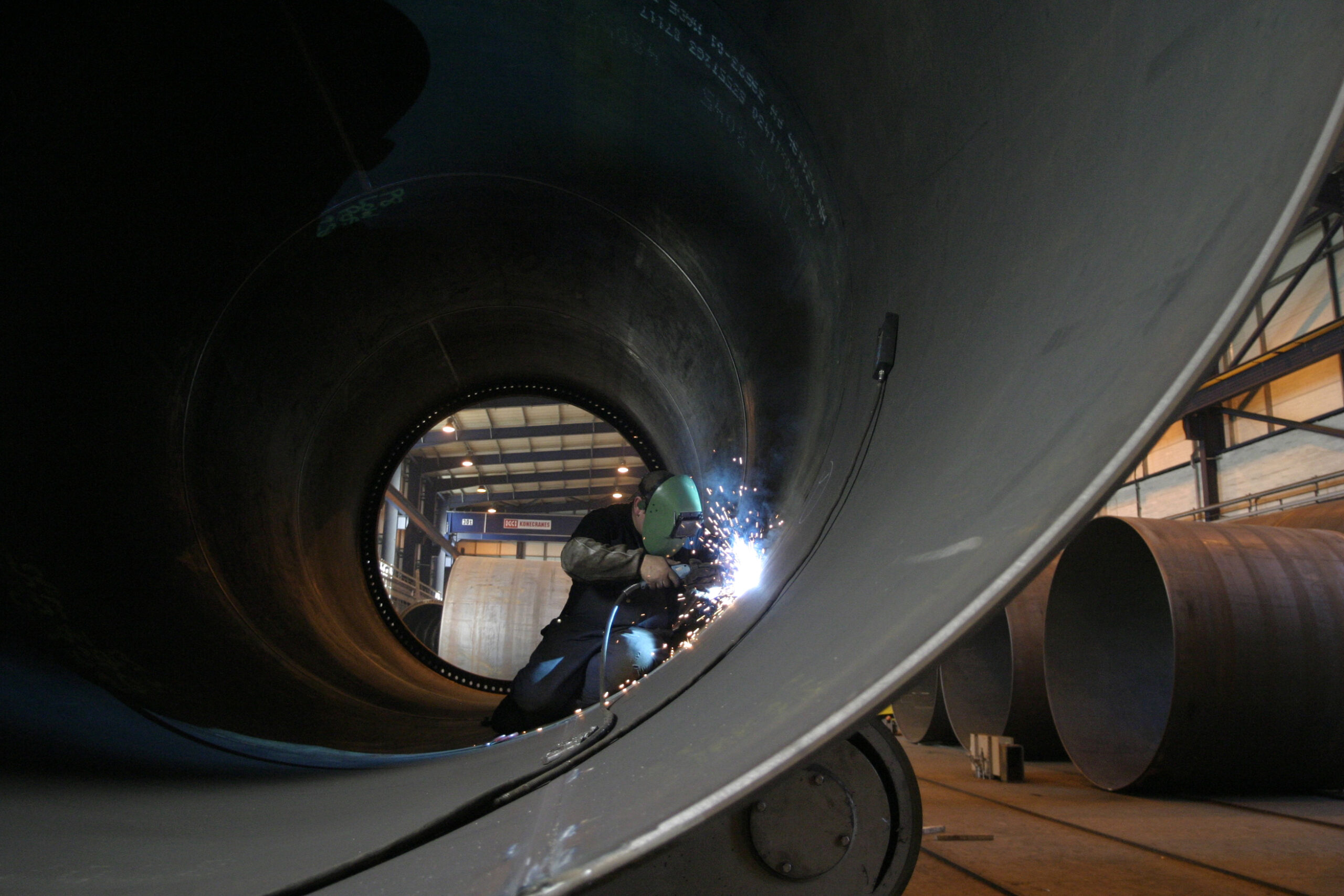 A person wearing protective gear welds the inside of a large cylindrical pipe in a factory.