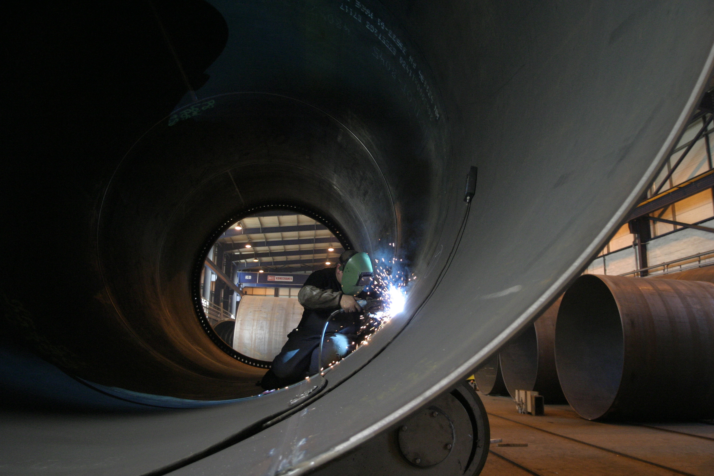 A person wearing protective gear welds the inside of a large cylindrical pipe in a factory.