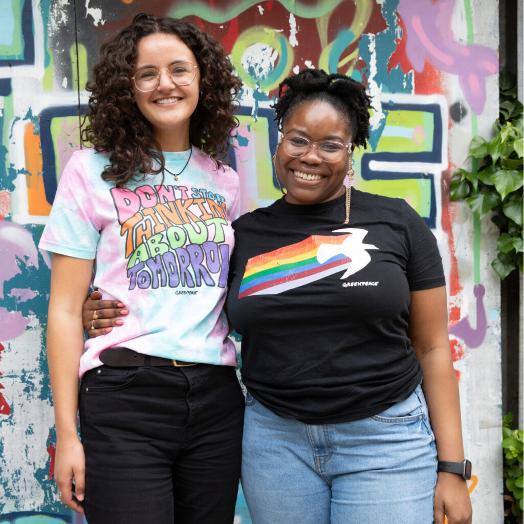 Two women in colourful greenpeace t-shirts hug and smile into the camera