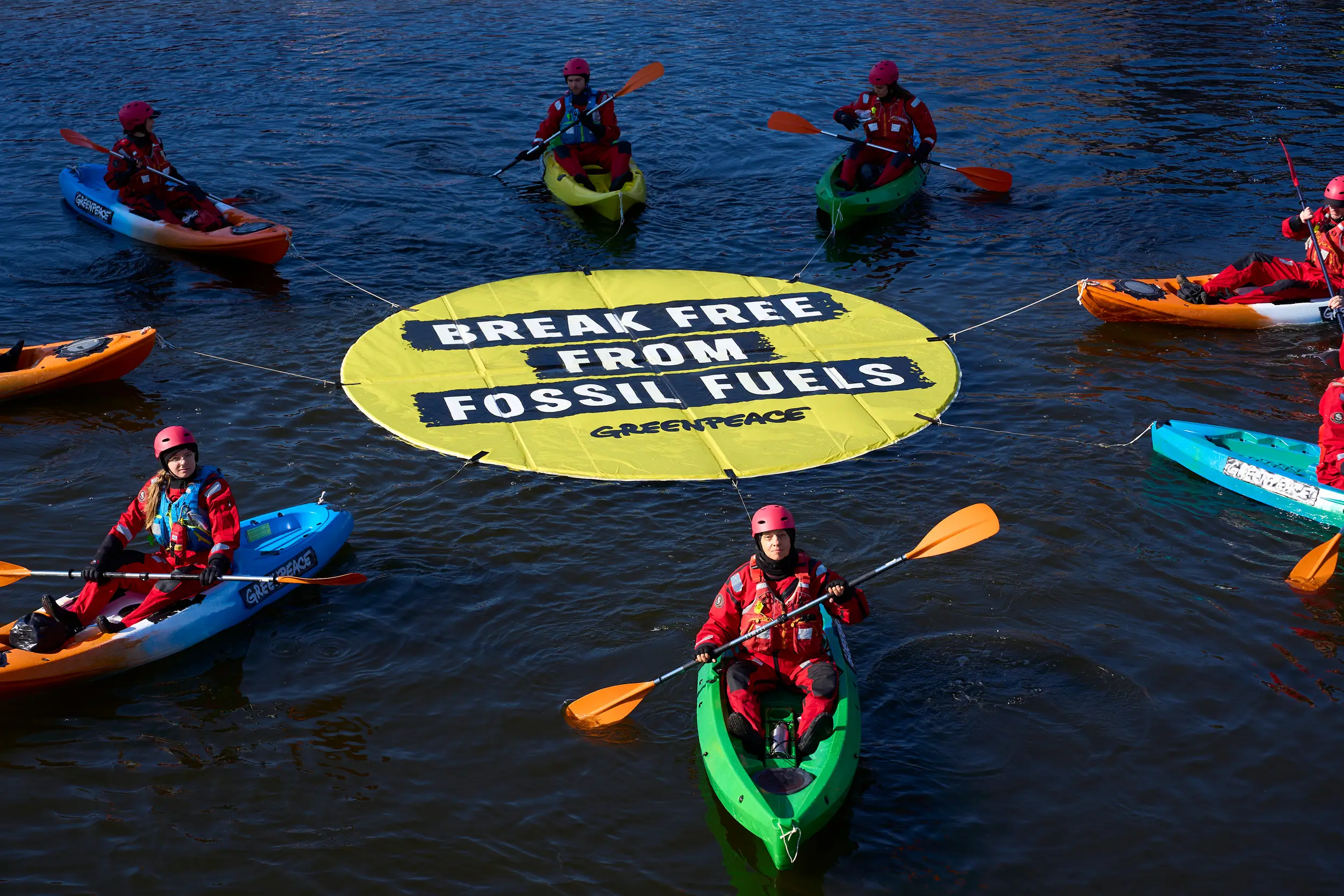 Kayakers in red suits and helmets surround a large yellow floating banner reading “Break Free From Fossil Fuels”